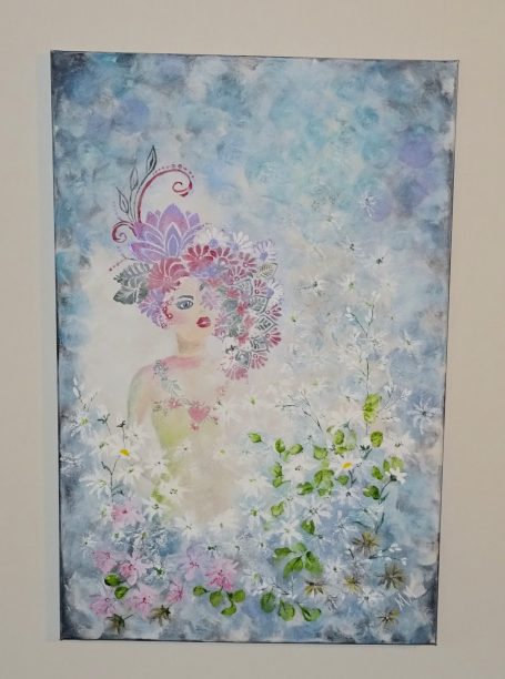 Kvinna med blommor i håret, omgiven av blå och vita blommor och mjuka färger.